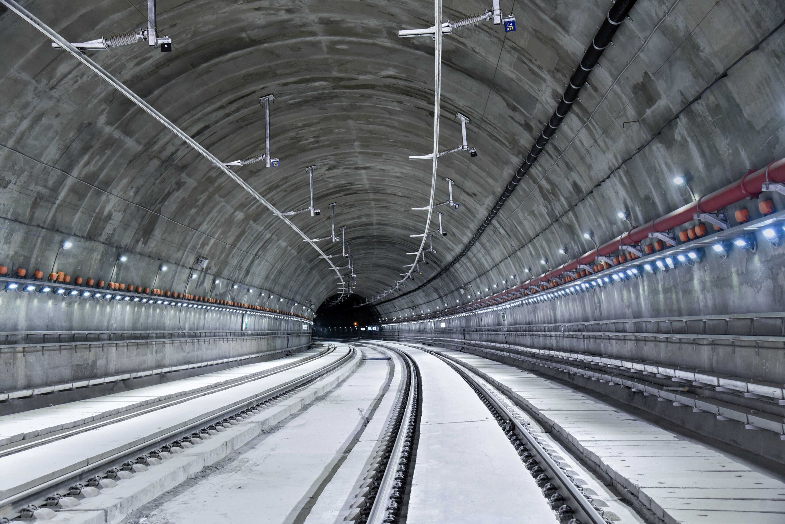 Ein gut beleuchteter, leerer unterirdischer Zugtunnel mit zwei Gleisen, die sich sanft nach links wölben, Betonwänden und einer mit Lampen und Kabeln ausgekleideten Decke. An der Decke eine Deckenstromschiene von Furrer+Frey