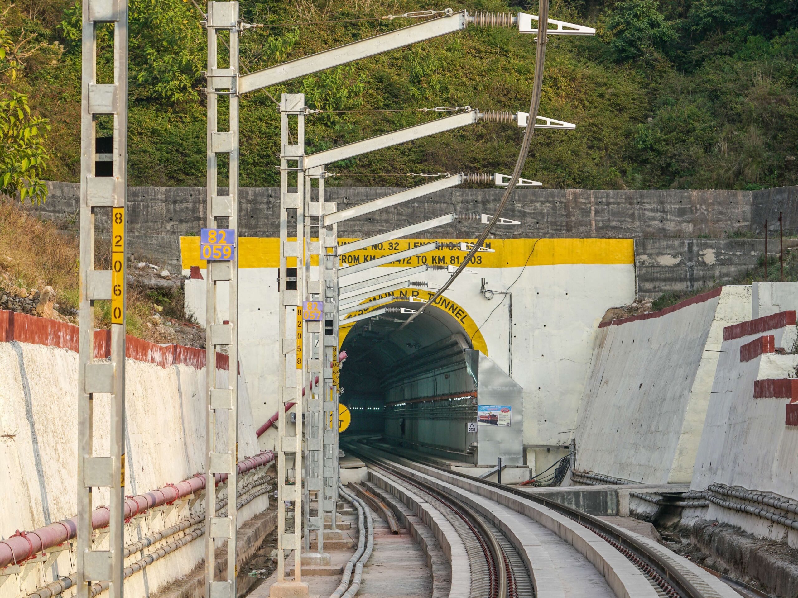Ein Bahngleis führt in einen betonierten Tunneleingang, der von Grünflächen und Stützmauern umgeben ist; gelbe Schilder und Fahrleitungsmasten säumen die Strecke. Es handelt sich hier um eine Deckenstromschiene von Furrer+Frey vor einem Tunnelportal