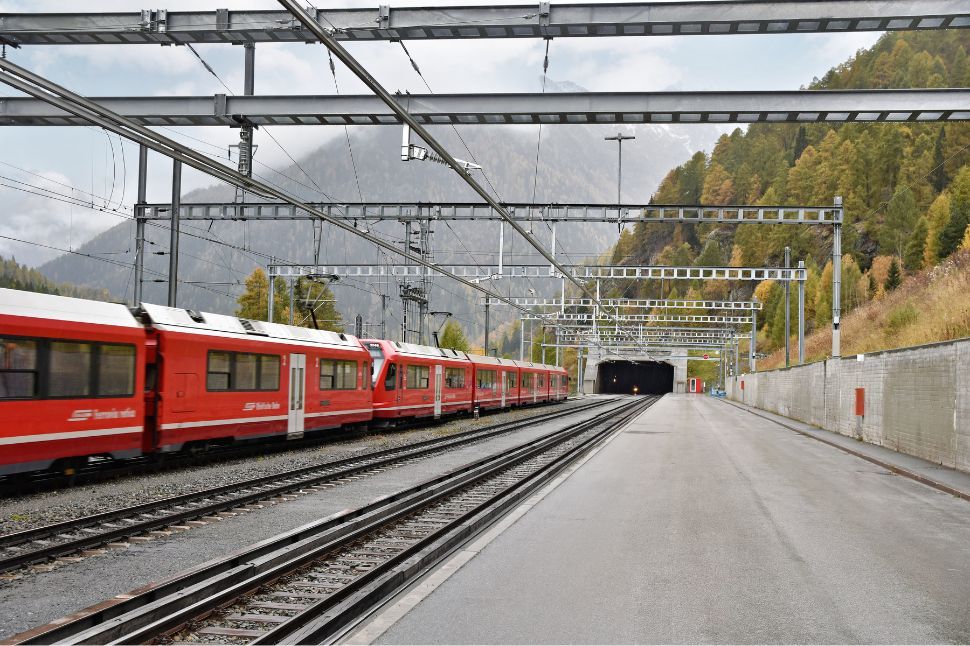 Ein roter Personenzug der Rhätischen Bahn fährt auf Eisenbahnschienen aus dem Vereinatunnel, mit Bergen und Bäumen im Hintergrund und Fahrleitungen über den Gleisen.