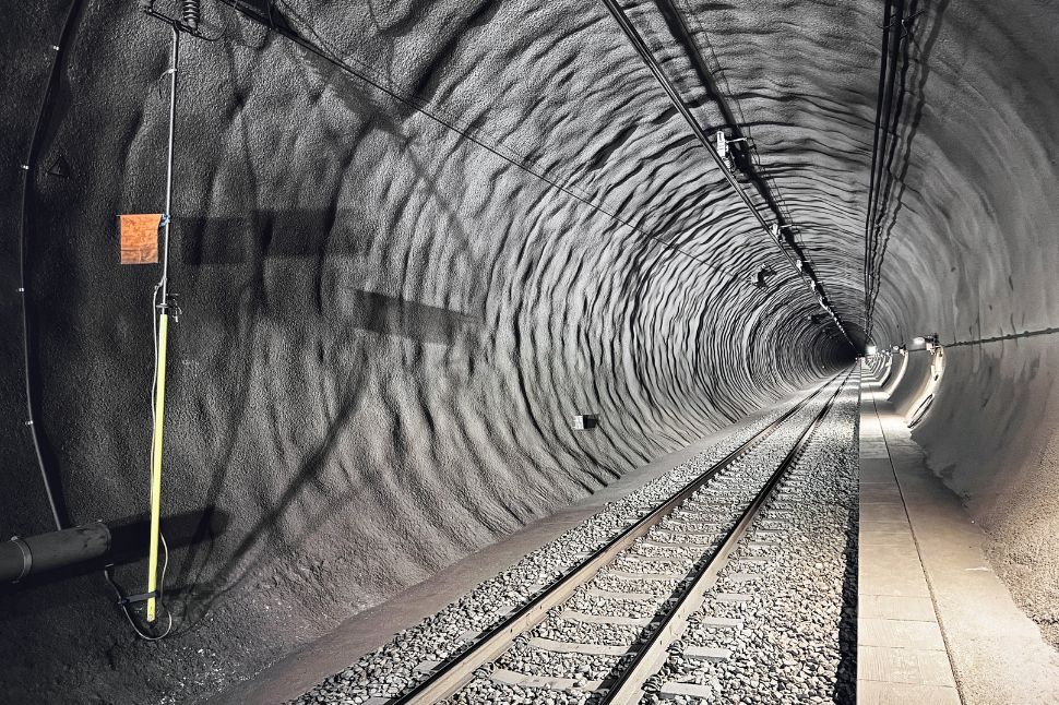 Ein gut beleuchteter Eisenbahntunnel mit strukturierten, gewellten Wänden. In der Mitte verlaufen Eisenbahnschienen, und auf der rechten Seite verläuft ein schmaler Gehweg. An den Wänden und der Decke des Tunnels sind elektrische Kabel und Geräte angebracht.